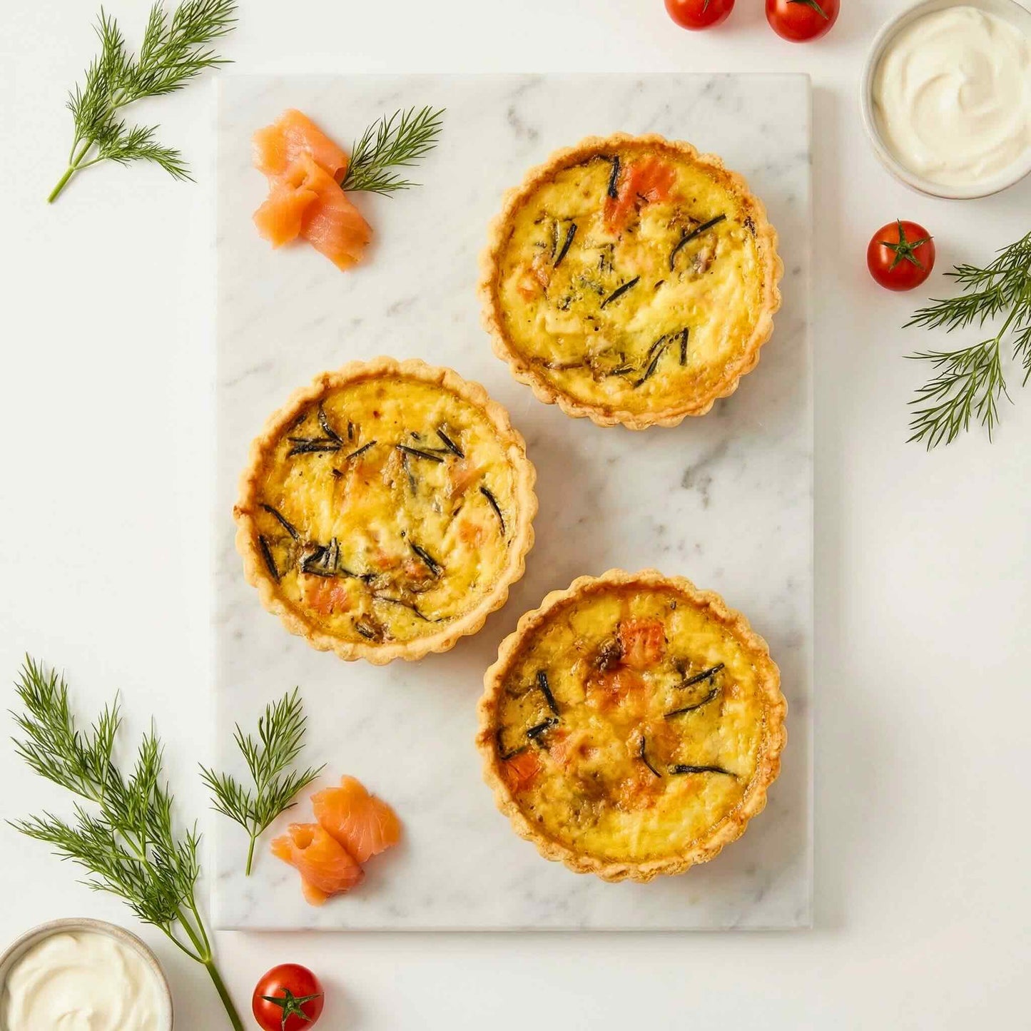 Overhead view of three single serve smoked salmon and caramelised onion quiches on white marble with fresh dill sprigs, smoked salmon pieces, cherry tomatoes and cream, 10cm individual size, Carpina Bakery Singapore