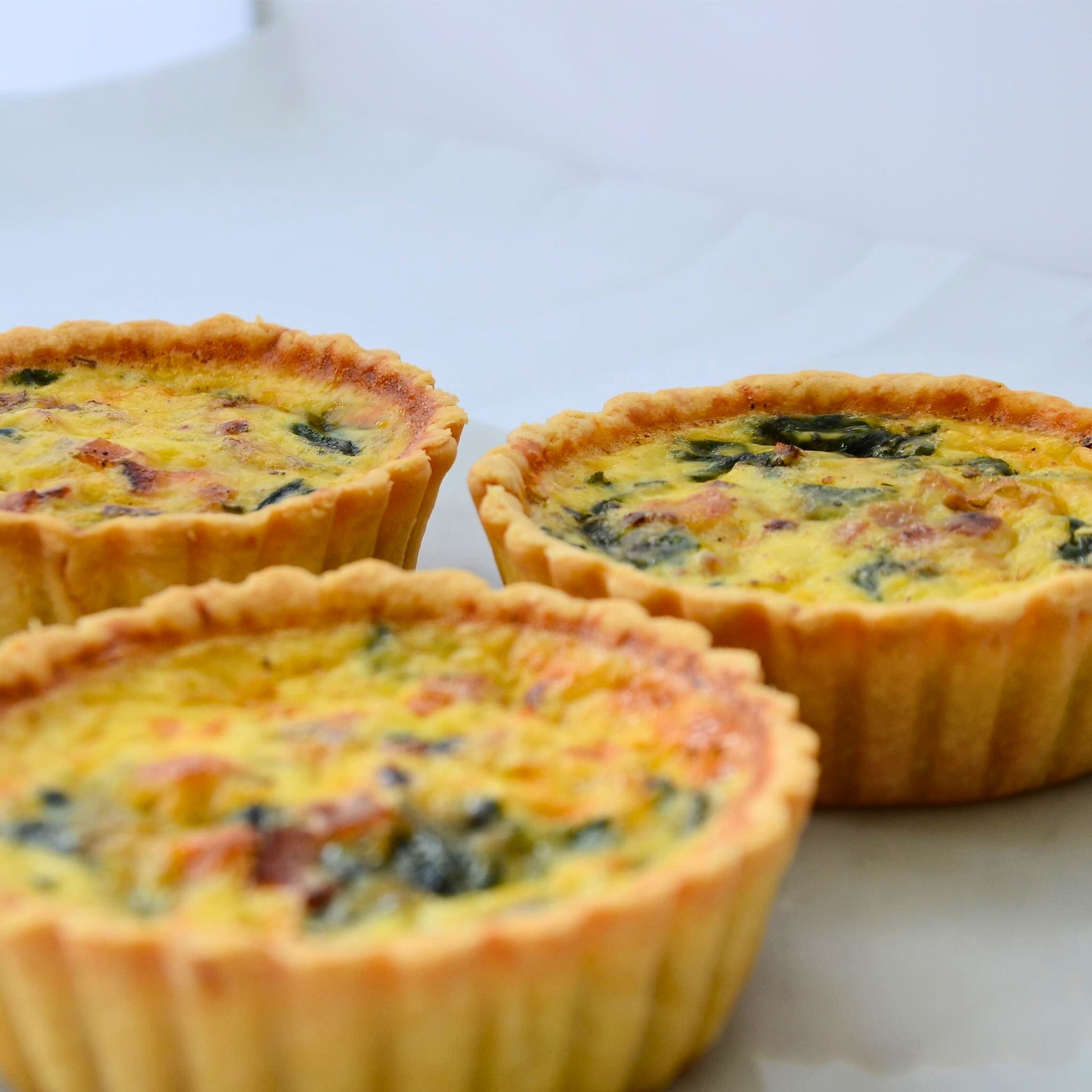 Three single serve Bacon Spinach quiches 10cm arranged on marble board showing golden buttery thin crust and creamy custard top with fresh spinach and crispy bacon Carpina Bakery Singapore