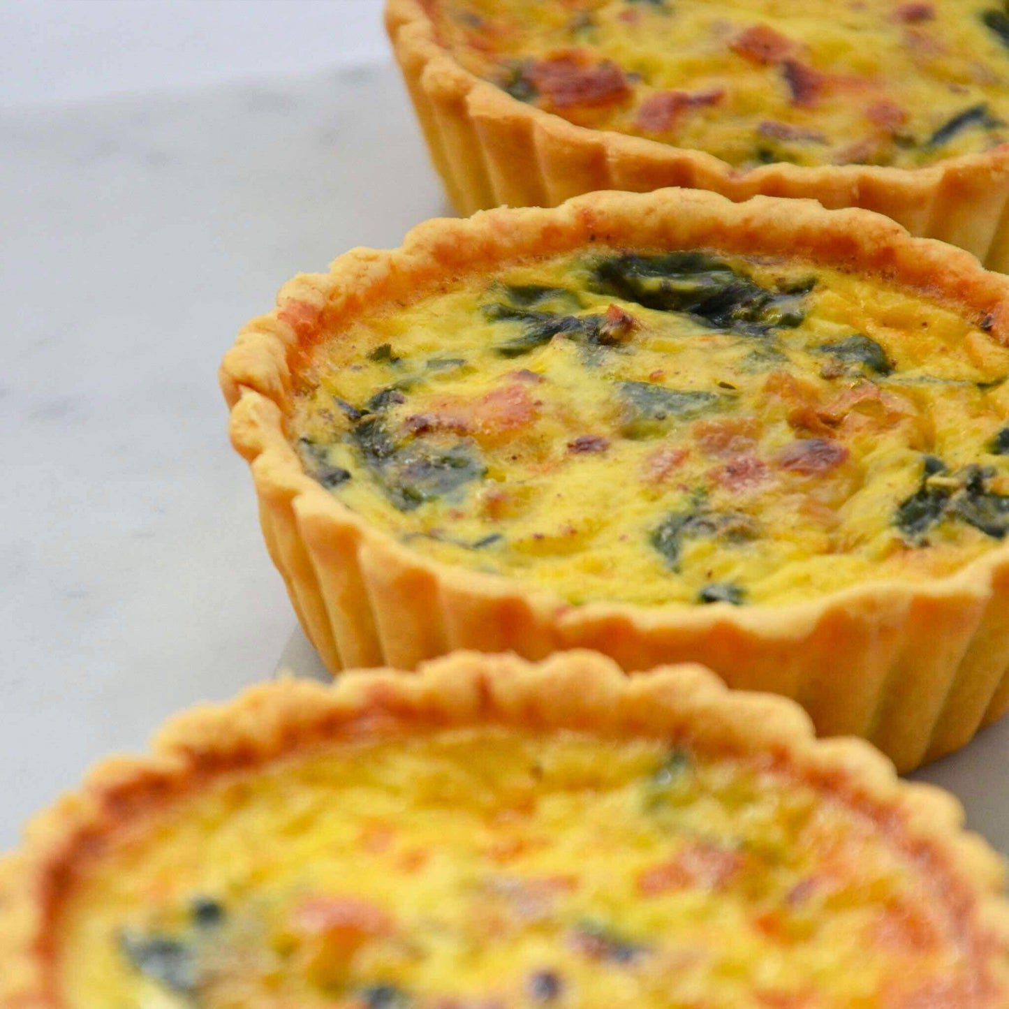 Single serve Bacon Spinach quiche 10cm whole quiche with golden buttery thin crust on marble surface fresh spinach leaf Carpina Bakery Singapore