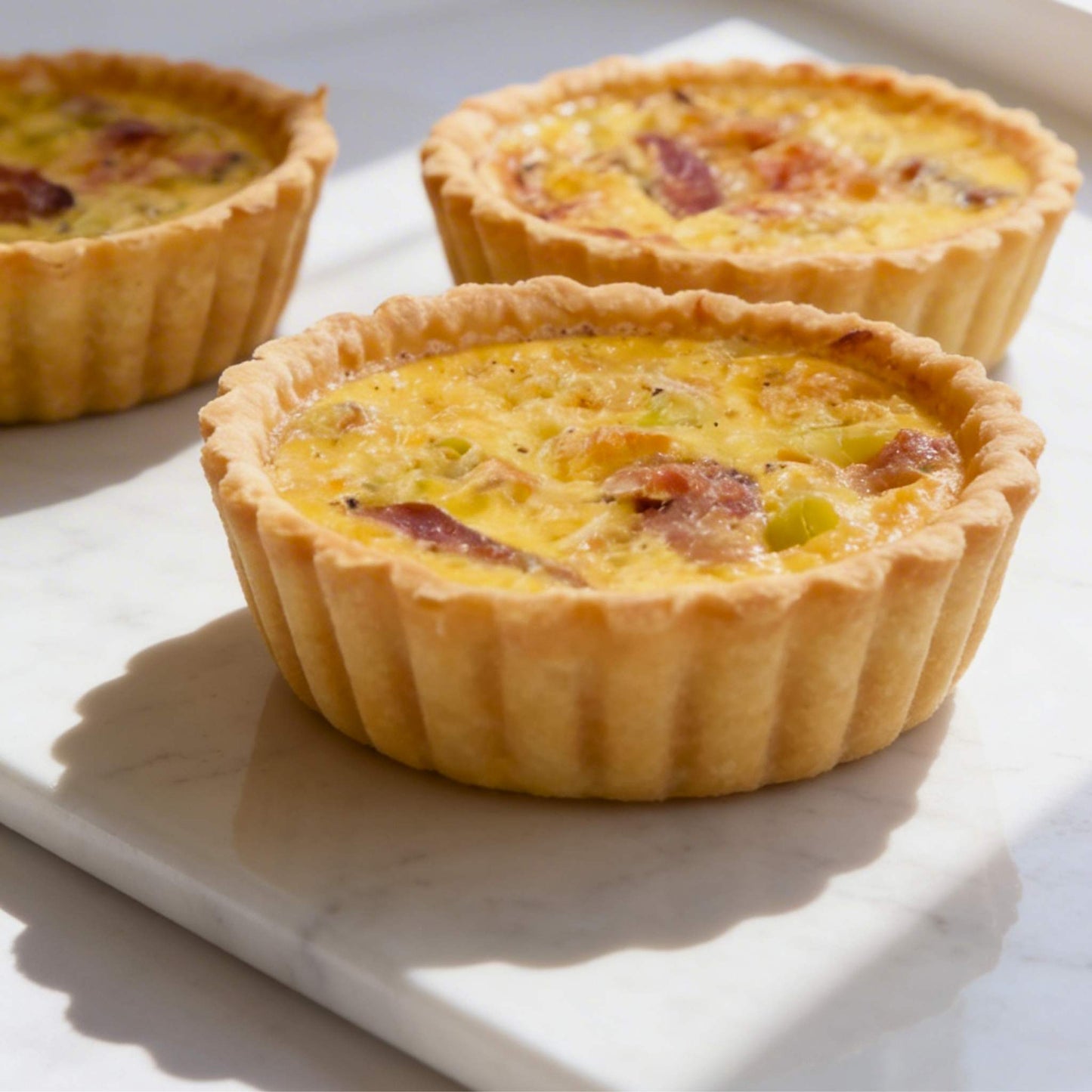 Close up of three single serve smoked duck and leek quiches on white marble showing flaky thin crust and creamy filling with smoked duck pieces and leeks, 10cm individual size, Carpina Bakery Singapore