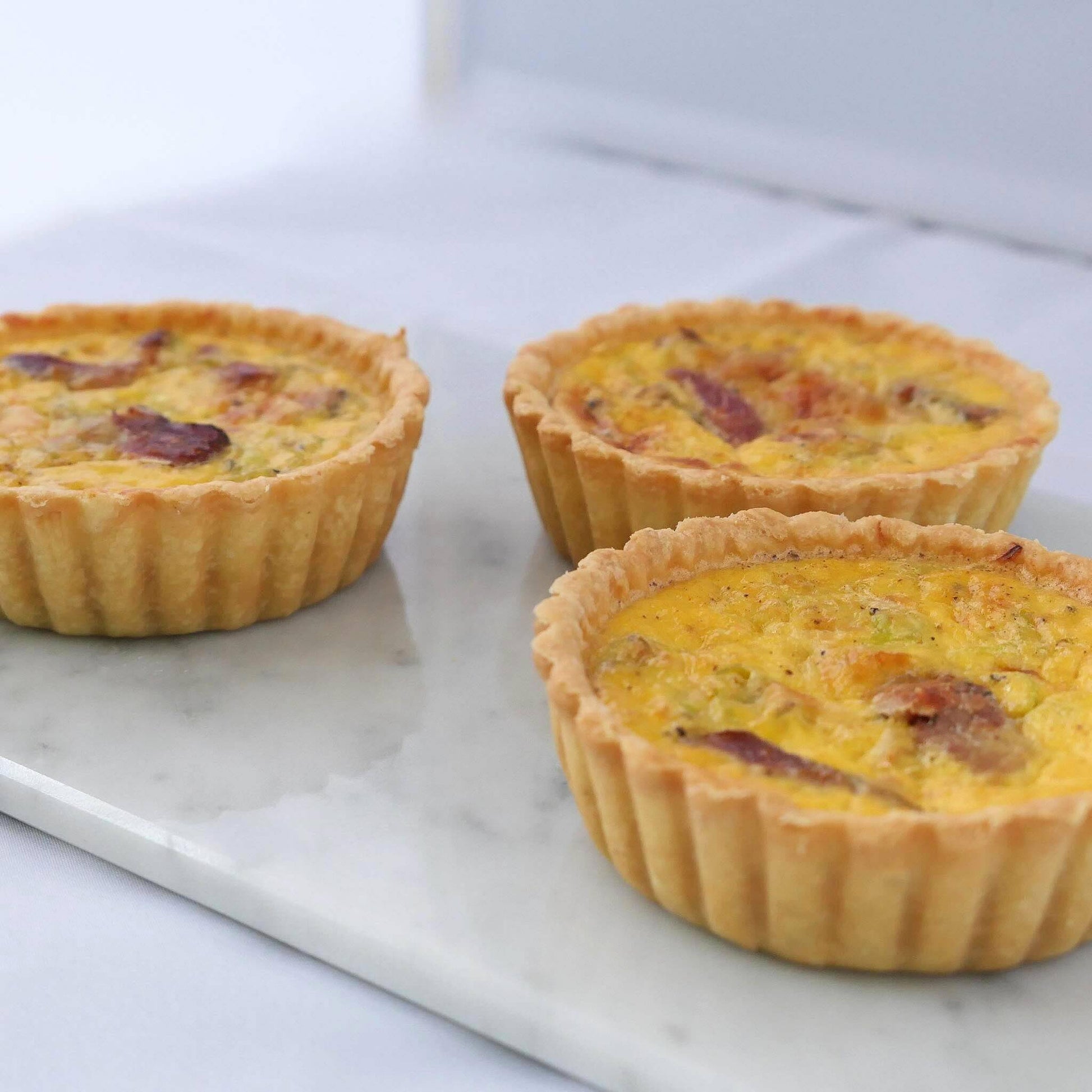 Three single serve smoked duck and leek quiches on white marble showing golden thin crust with smoked duck pieces and leeks in creamy filling, 10cm individual size, Carpina Bakery Singapore
