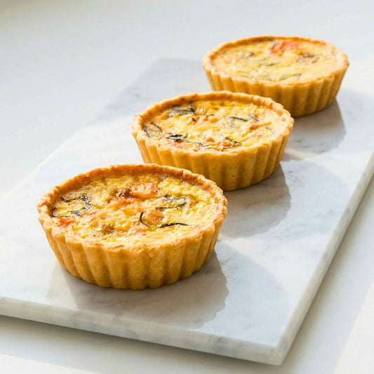 Three single serve smoked salmon and caramelised onion quiches on white marble showing golden thin crust with rosemary and smoked salmon pieces in creamy filling, 10cm individual size, Carpina Bakery Singapore