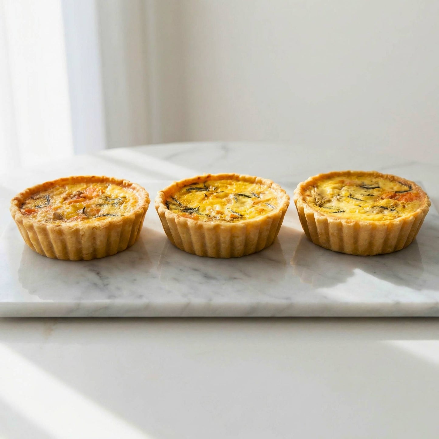 Three single serve smoked salmon and caramelised onion quiches arranged in a row on white marble with natural light, 10cm individual size, Carpina Bakery Singapore