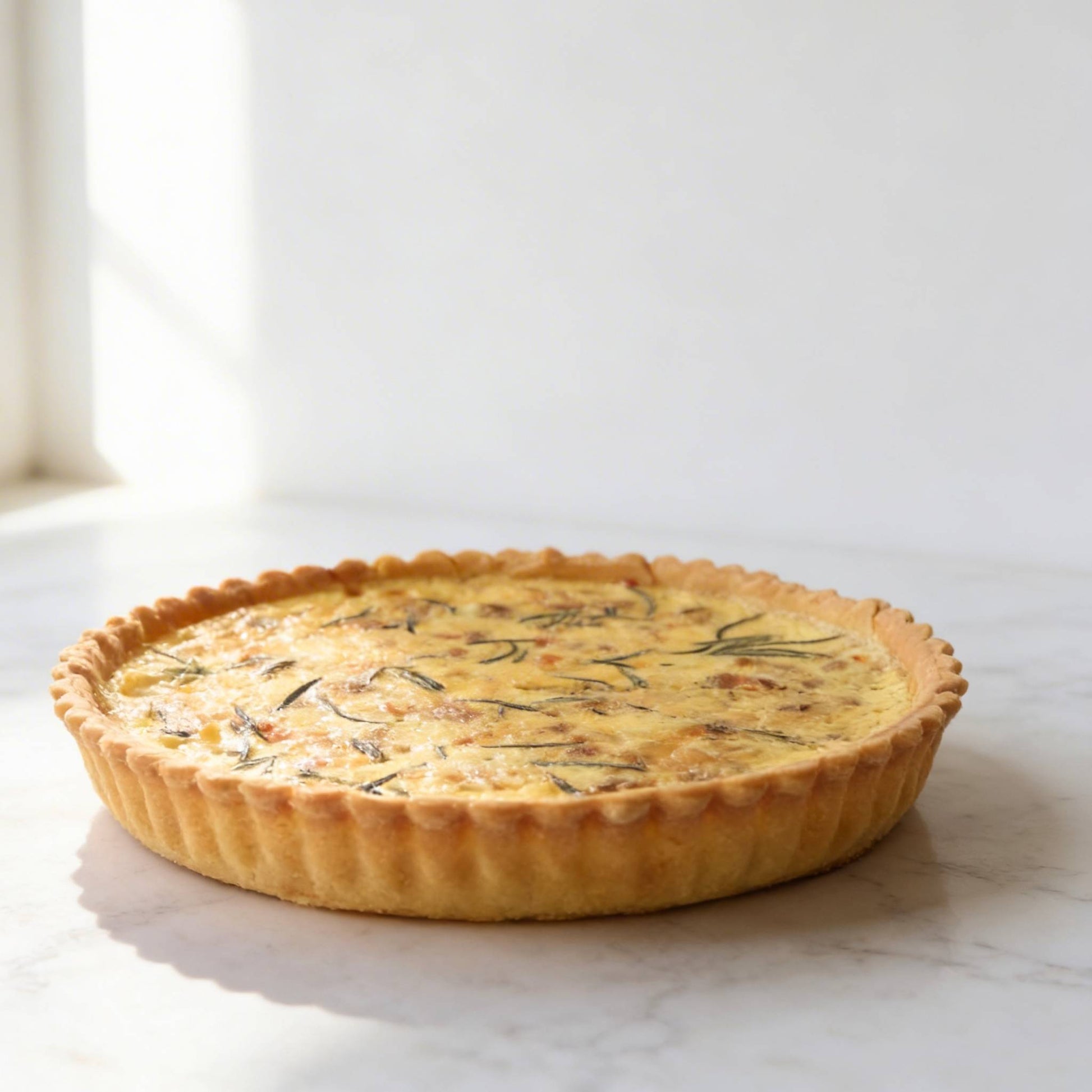 Side view of whole Smoked Salmon and Caramelised Onion Quiche on white marble showing fluted golden thin crust and creamy filling, 24cm family size, Carpina Bakery Singapore